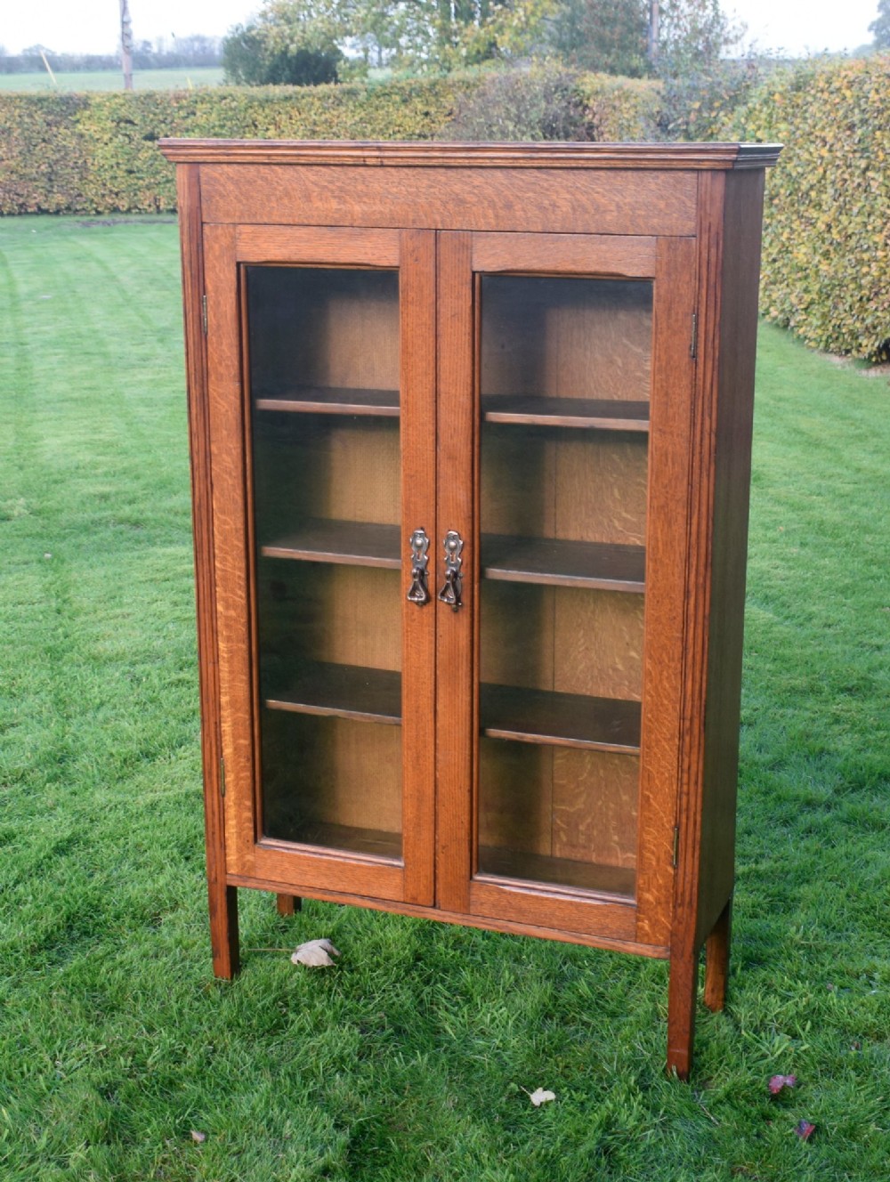 An early twentieth-century warm brown oak bookcase featuring twin glazed doors, brass hardware, and three internal shelves. View VI