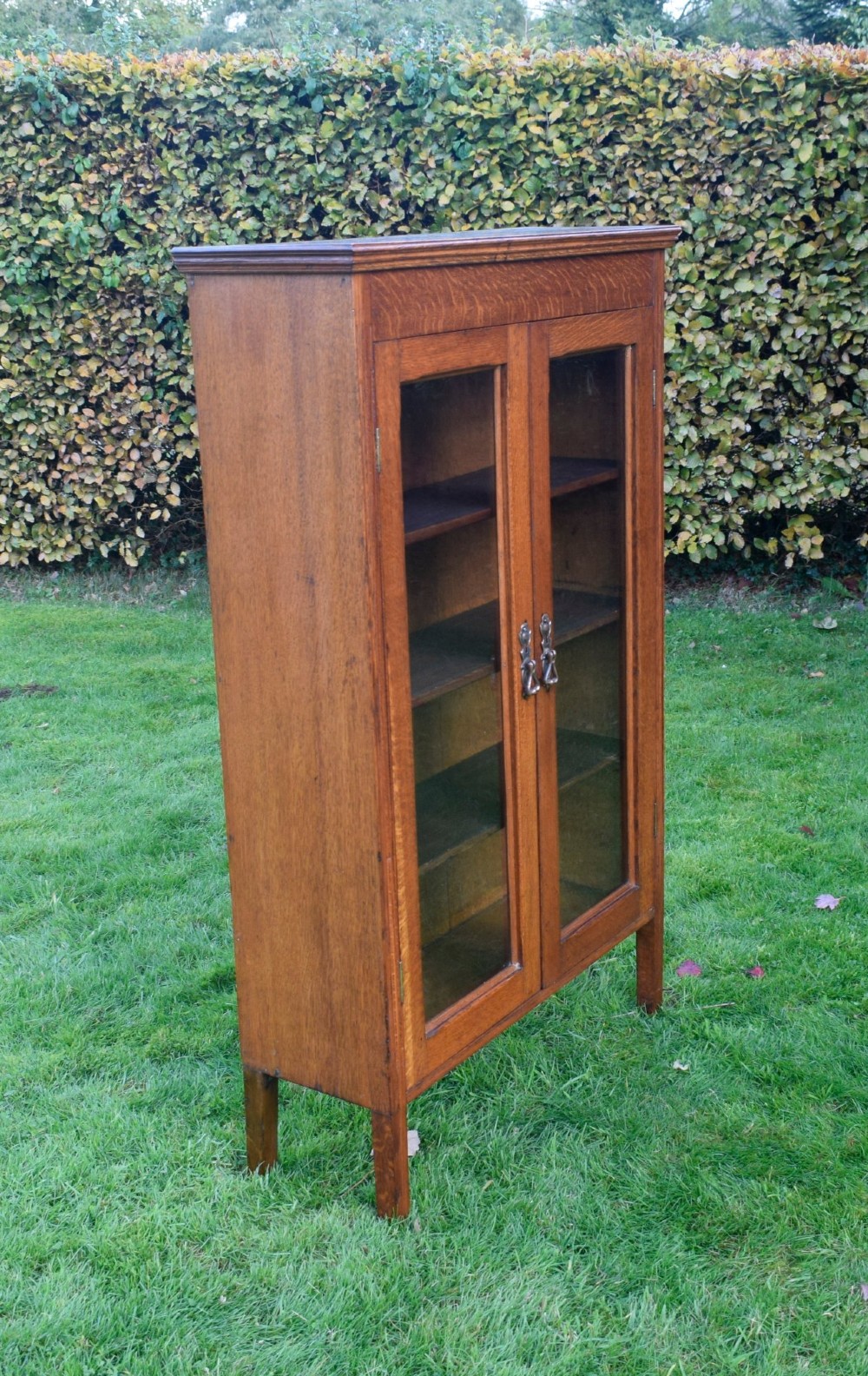 An early twentieth-century warm brown oak bookcase featuring twin glazed doors, brass hardware, and three internal shelves. View VIII