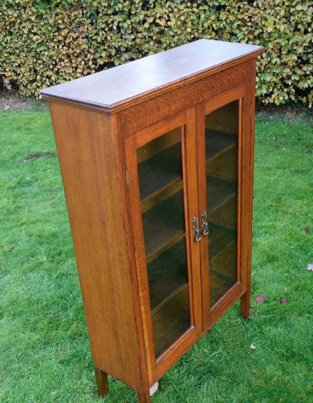 An early twentieth-century warm brown oak bookcase featuring twin glazed doors, brass hardware, and three internal shelves. View IX