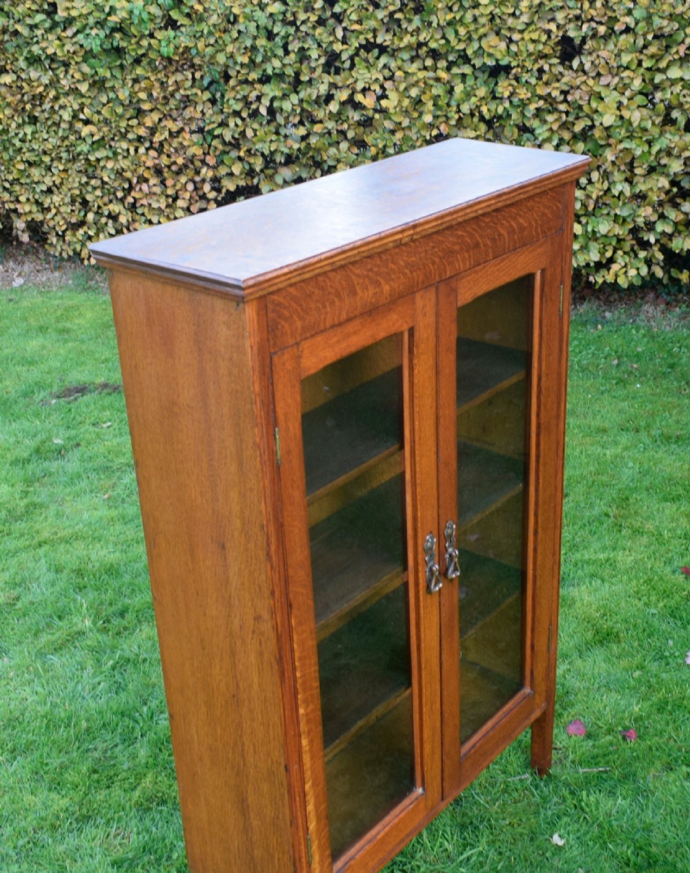 An early twentieth-century warm brown oak bookcase featuring twin glazed doors, brass hardware, and three internal shelves. View X