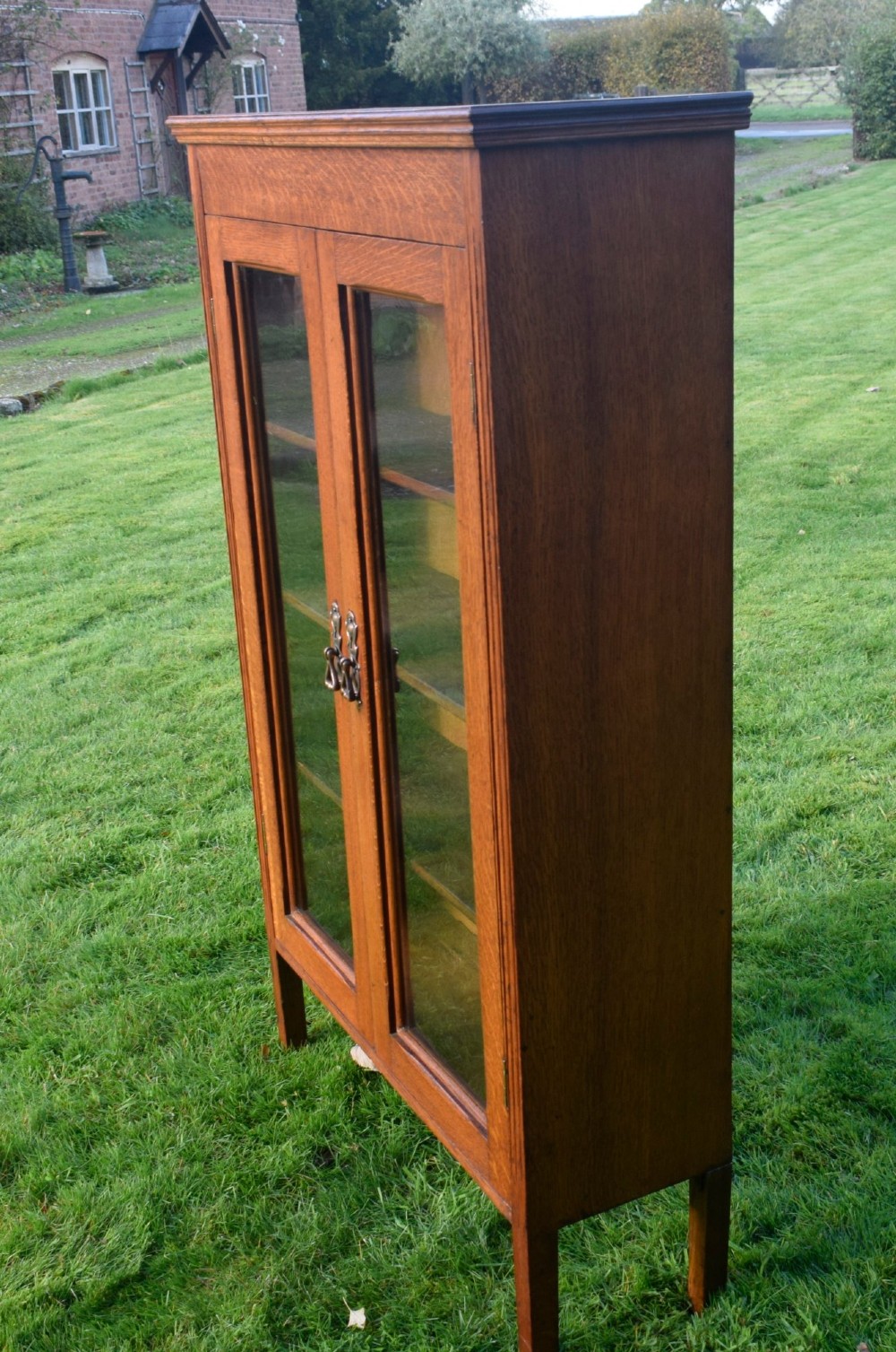 An early twentieth-century warm brown oak bookcase featuring twin glazed doors, brass hardware, and three internal shelves. View XIII