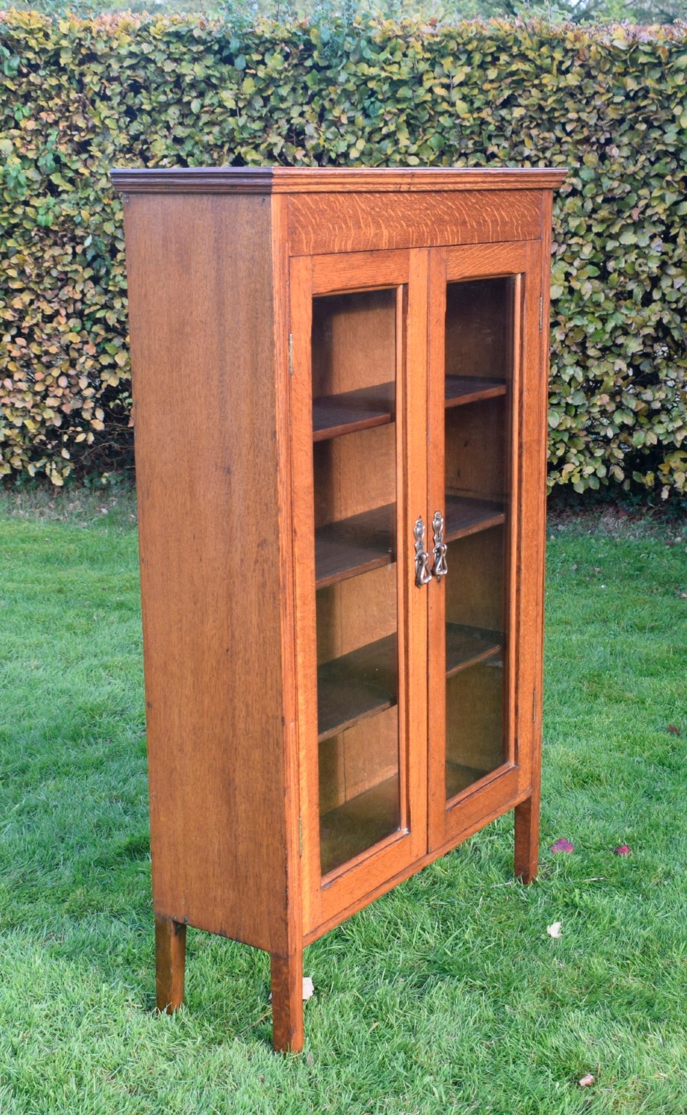 An early twentieth-century warm brown oak bookcase featuring twin glazed doors, brass hardware, and three internal shelves. View XV