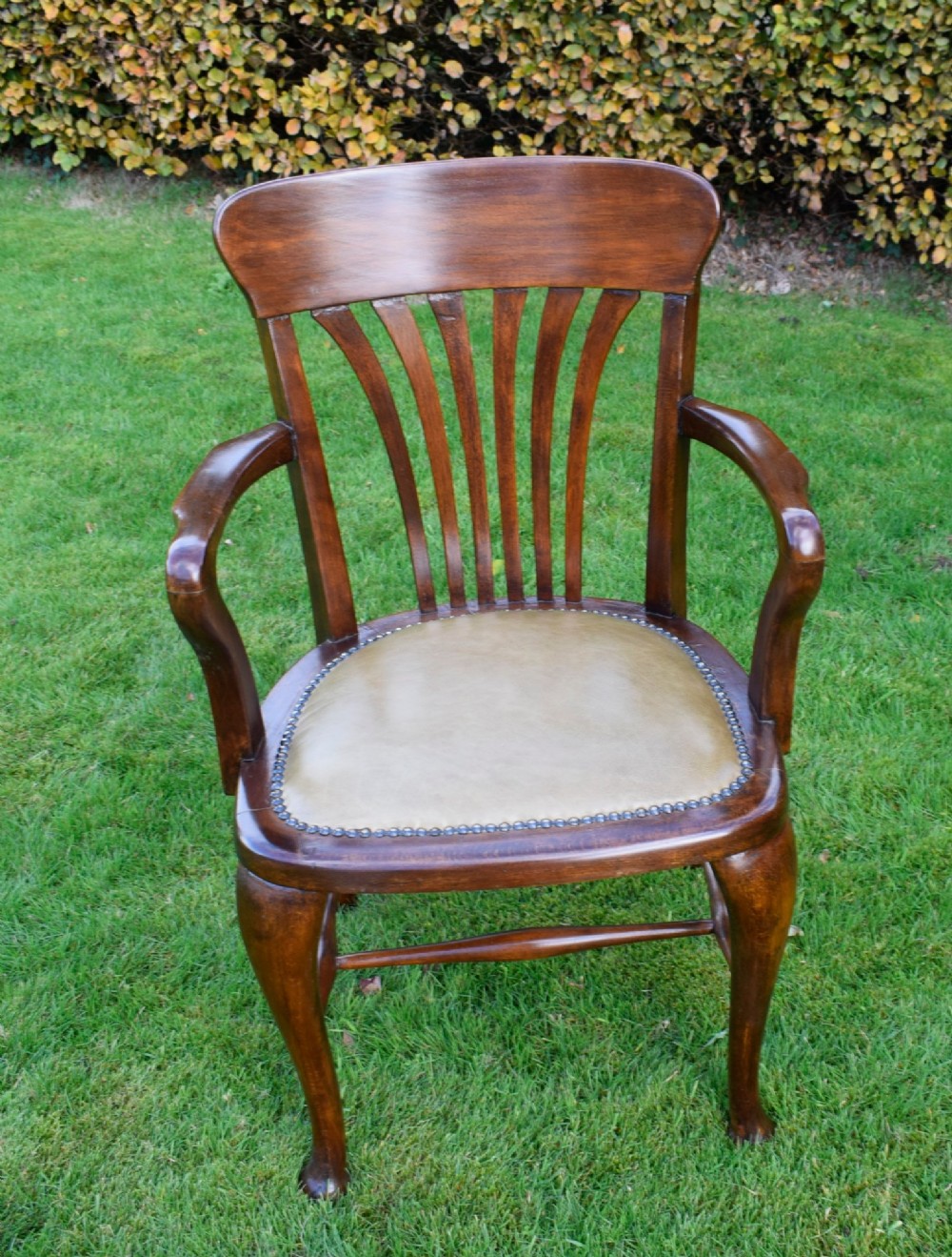 A late Victorian wooden desk armchair with a curved slatted backrest, curved arms, and a tan leather seat trimmed with studding. View X