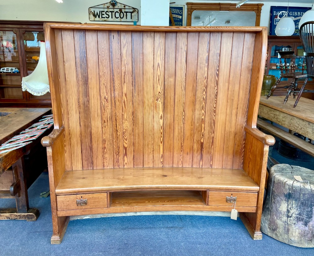 A high-backed pine settle featuring vertical tongue-and-groove panelling, a gently curved seat, two base drawers with brass handles, and an open central shelf.