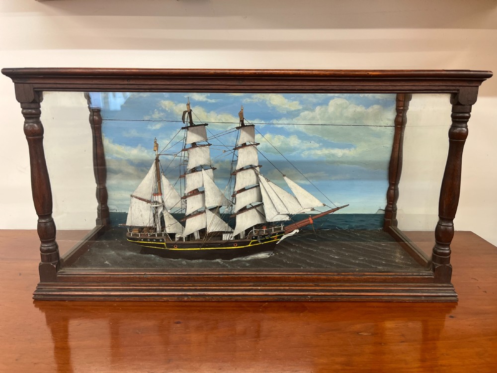 A 19th-century English ship diorama featuring a detailed three-masted sailing vessel set against a painted ocean background inside a glazed wooden case with turned corner posts.