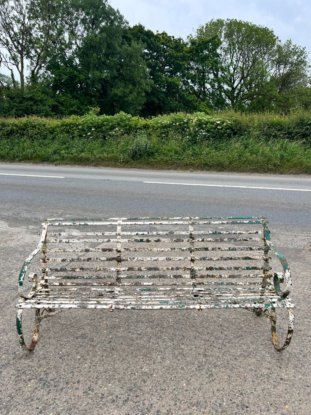 A Victorian wrought iron garden bench featuring horizontal metal strapwork, curved scroll armrests, and a distressed green and white painted finish.