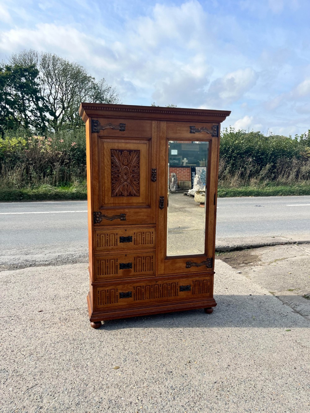 A turn-of-the-century Edwardian oak gentlemans wardrobe featuring a full-length mirrored door, relief-carved central panel, three carved front drawers, and iron strap hinges.