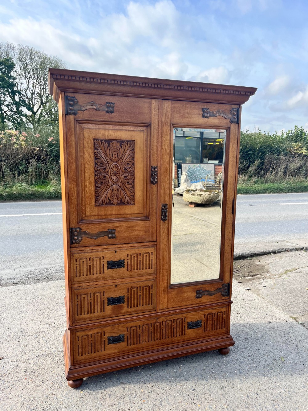 A turn-of-the-century Edwardian oak gentlemans wardrobe featuring a full-length mirrored door, relief-carved central panel, three carved front drawers, and iron strap hinges. View II