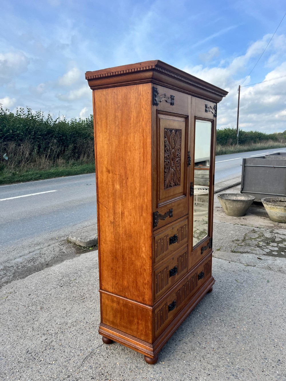 A turn-of-the-century Edwardian oak gentlemans wardrobe featuring a full-length mirrored door, relief-carved central panel, three carved front drawers, and iron strap hinges. View XI