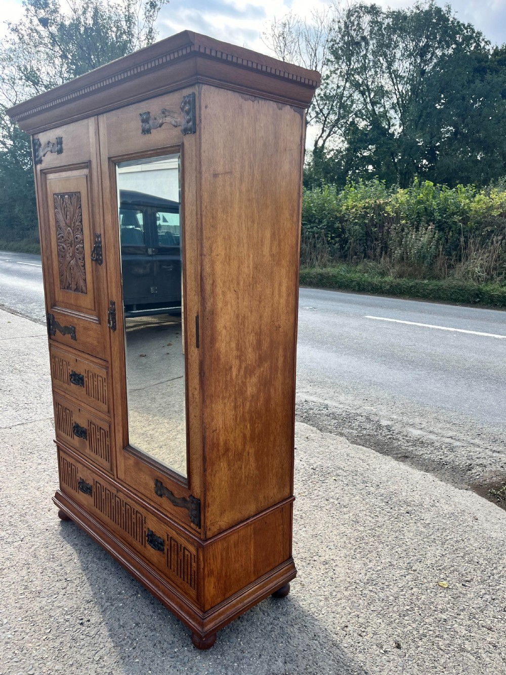 A turn-of-the-century Edwardian oak gentlemans wardrobe featuring a full-length mirrored door, relief-carved central panel, three carved front drawers, and iron strap hinges. View XIII