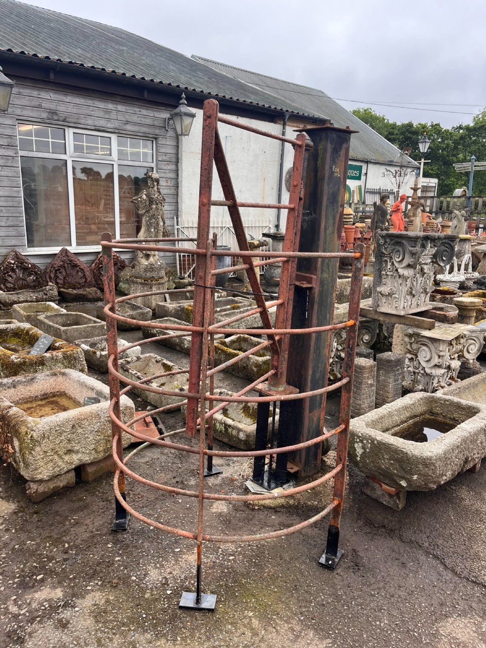 An antique wrought iron kissing gate featuring a curved enclosure and weathered red paint, mounted on a central iron post. View X