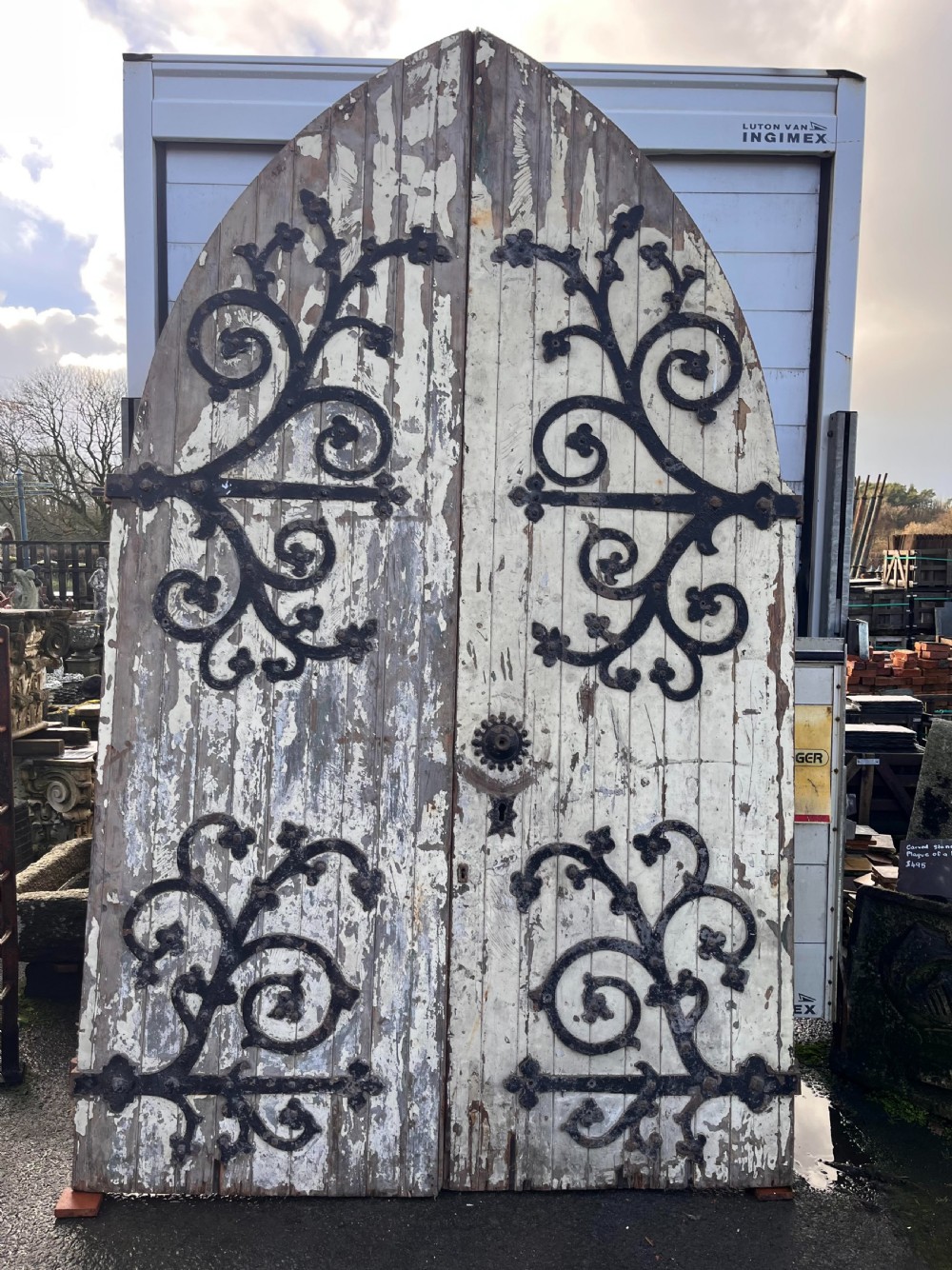 A pair of large, arched Victorian pine chapel doors featuring distressed white paint and elaborate black wrought iron scrollwork strap hinges.