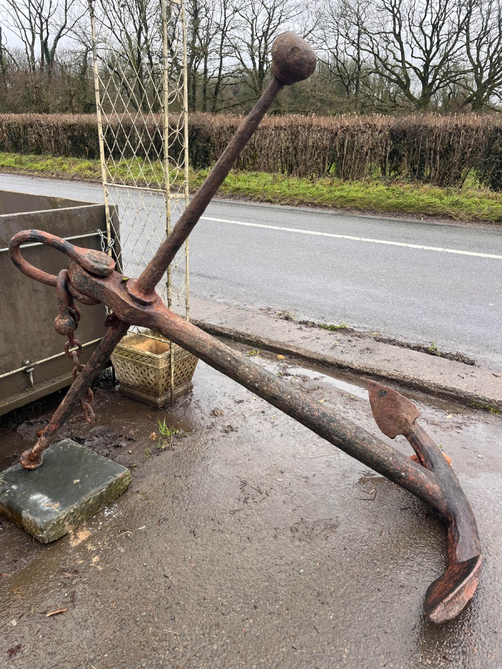 A large 18th-century hand-forged wrought iron ship anchor with a weathered rust finish resting outdoors on gravel.