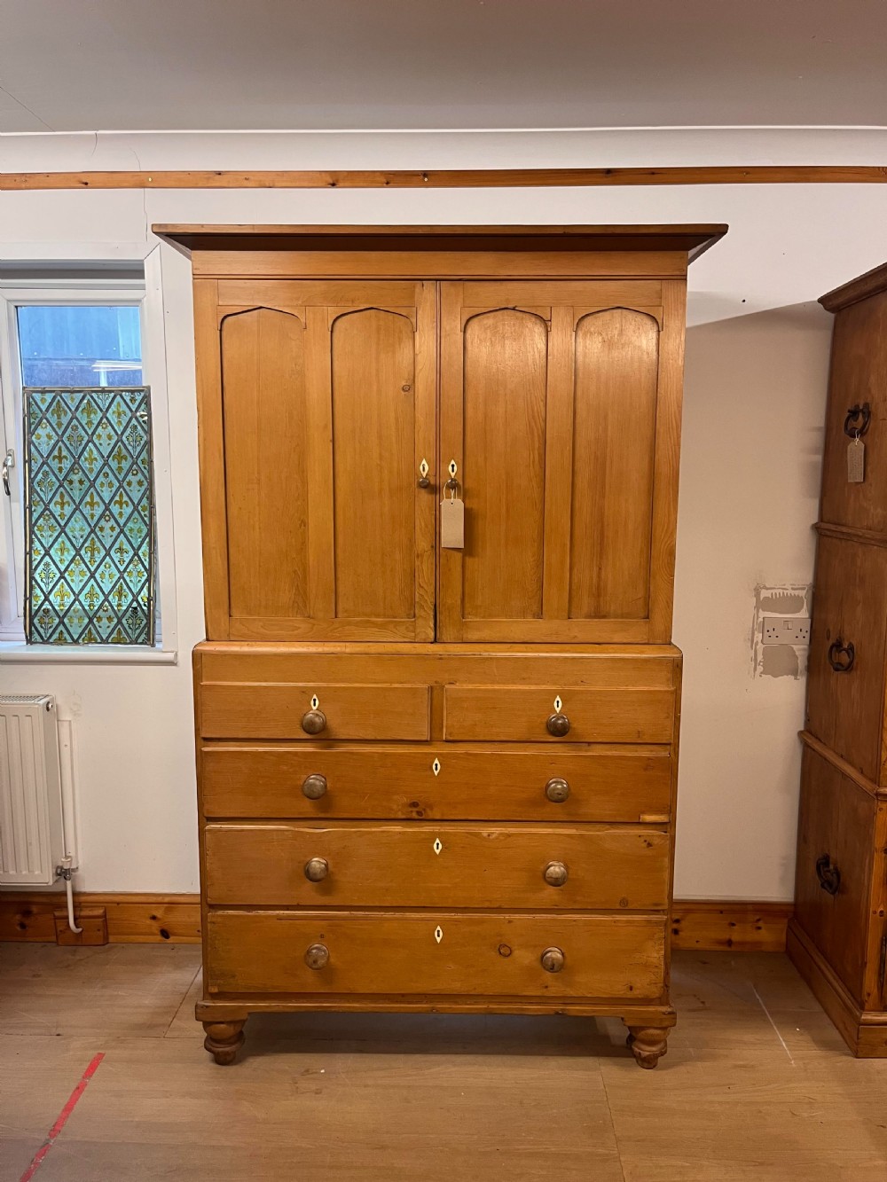A Victorian pine linen press featuring a two-door upper cupboard with arched fielded panels over a chest of drawers with turned wooden knobs and feet.
