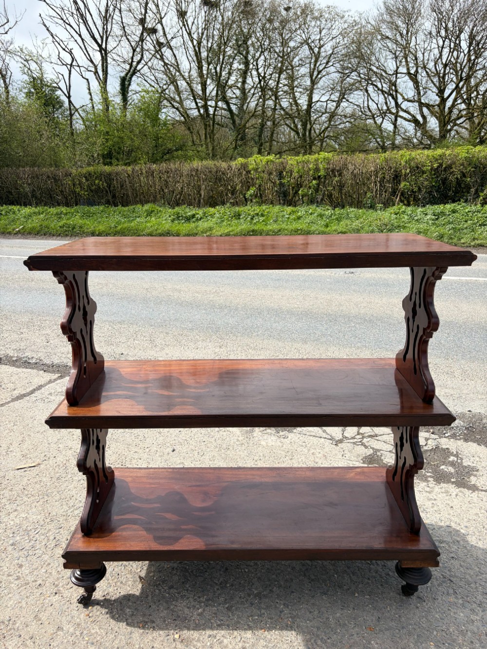 A Victorian mahogany three-tier buffet featuring carved pierced side supports, open rectangular shelves, and turned feet resting on original castors.