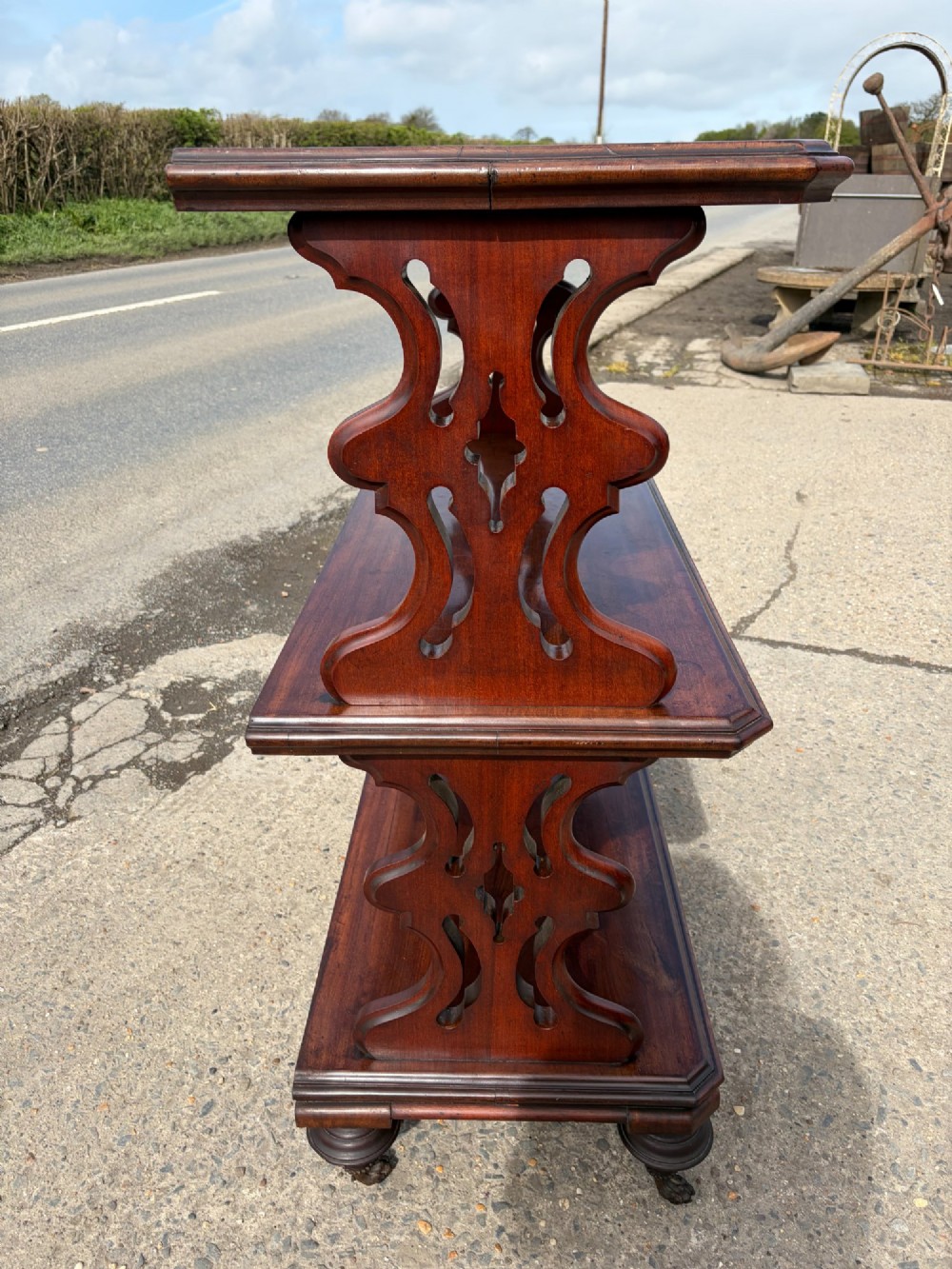 A Victorian mahogany three-tier buffet featuring carved pierced side supports, open rectangular shelves, and turned feet resting on original castors. View II