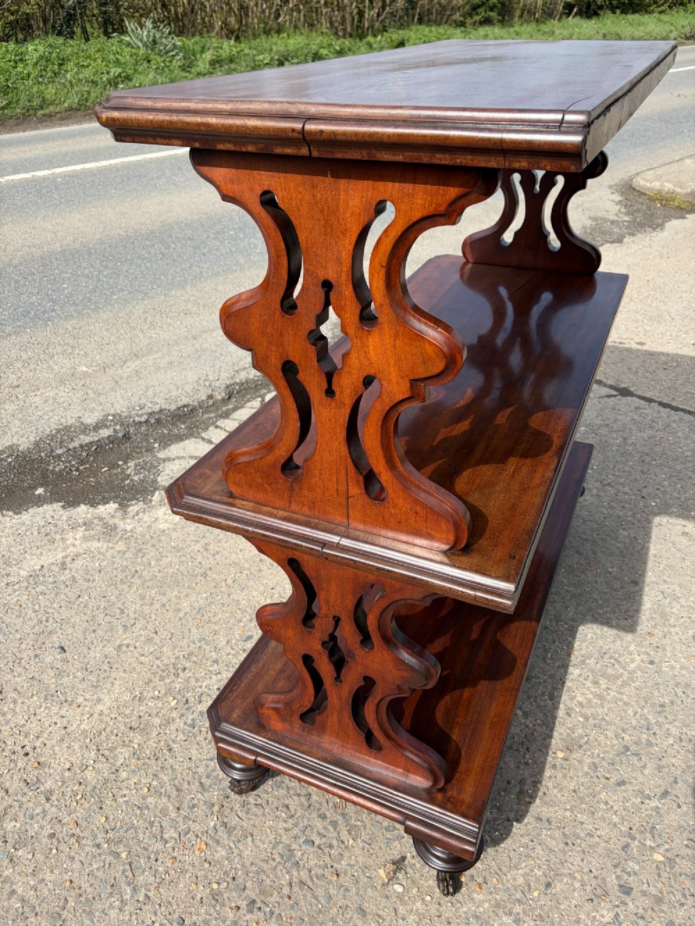 A Victorian mahogany three-tier buffet featuring carved pierced side supports, open rectangular shelves, and turned feet resting on original castors. View XII
