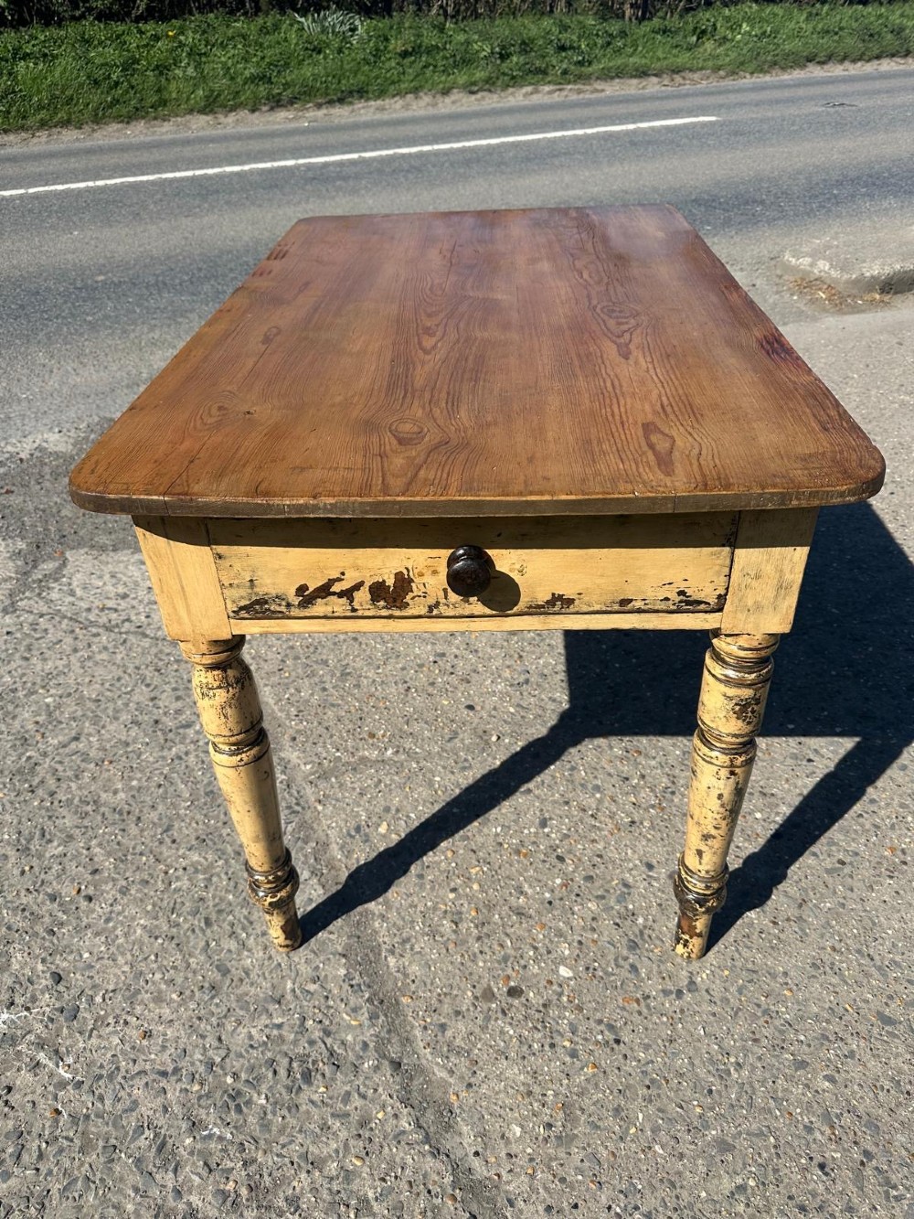 A Victorian pine table featuring a natural wood top, a single drawer with a round wooden knob, and turned yellow painted legs showing light surface wear.
