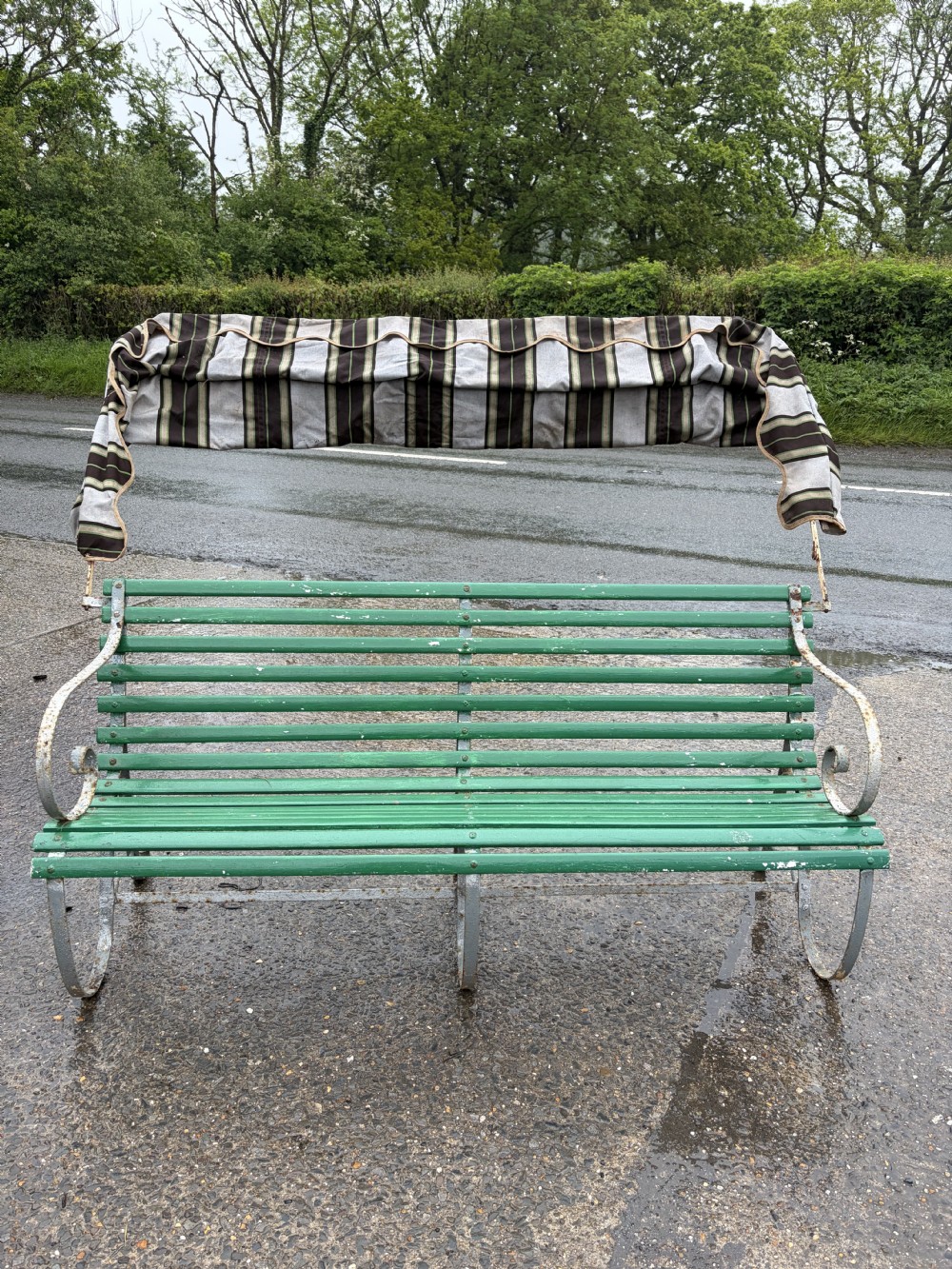 A Victorian wrought iron garden bench painted green with a slatted seat and an overhead striped canvas canopy. View V