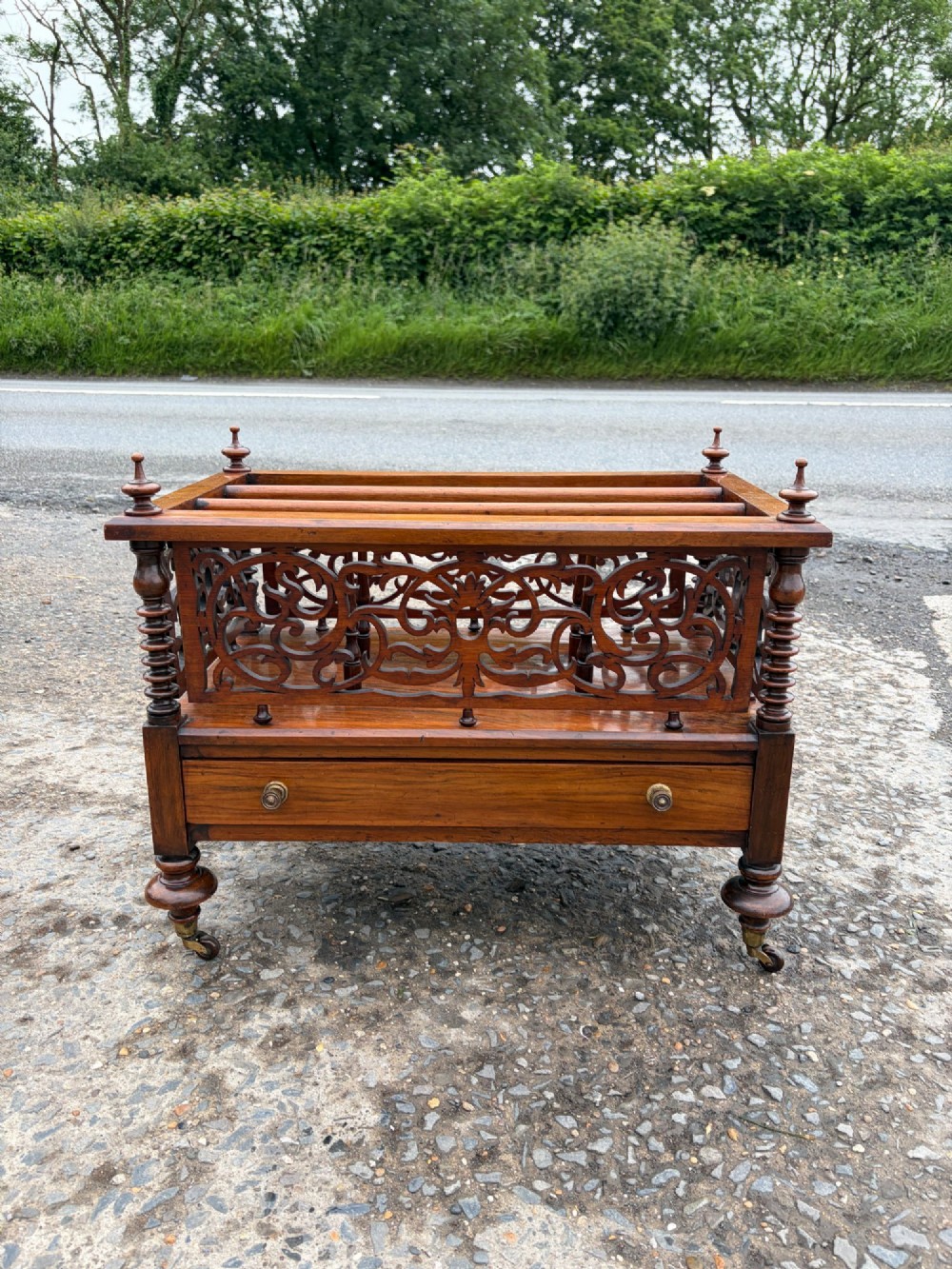 A Victorian walnut canterbury with pierced fretwork dividers, turned corner posts, finials, a lower drawer, and brass castors.