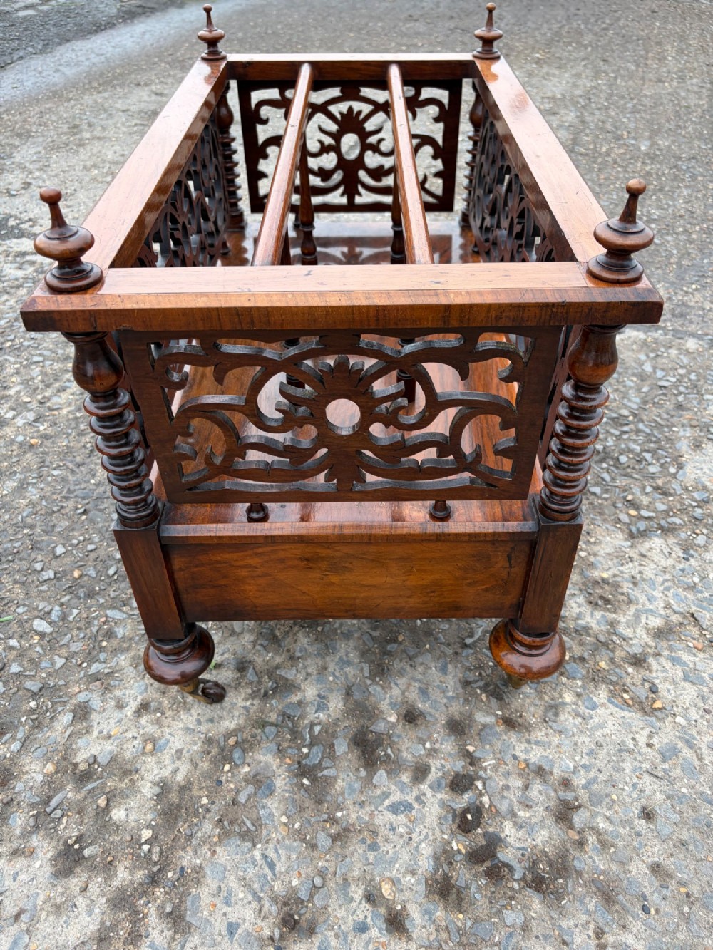 A Victorian walnut canterbury with pierced fretwork dividers, turned corner posts, finials, a lower drawer, and brass castors. View II
