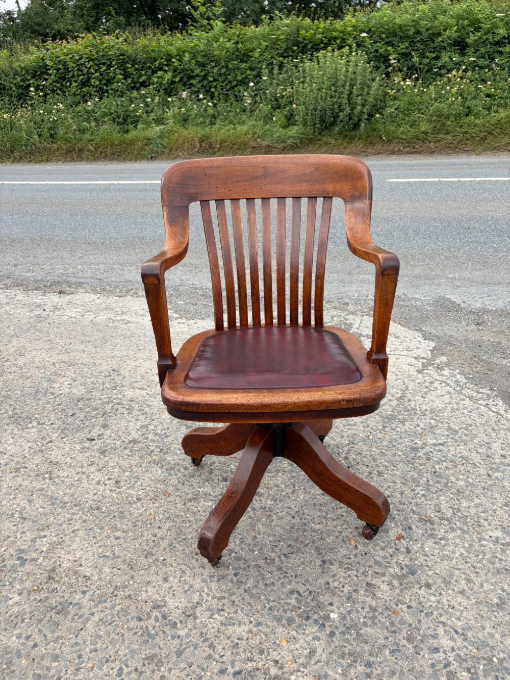 A 1920s solid oak swivel desk chair featuring a slatted backrest, integrated armrests, a reddish-brown leather seat cushion, and a four-star base on castors.