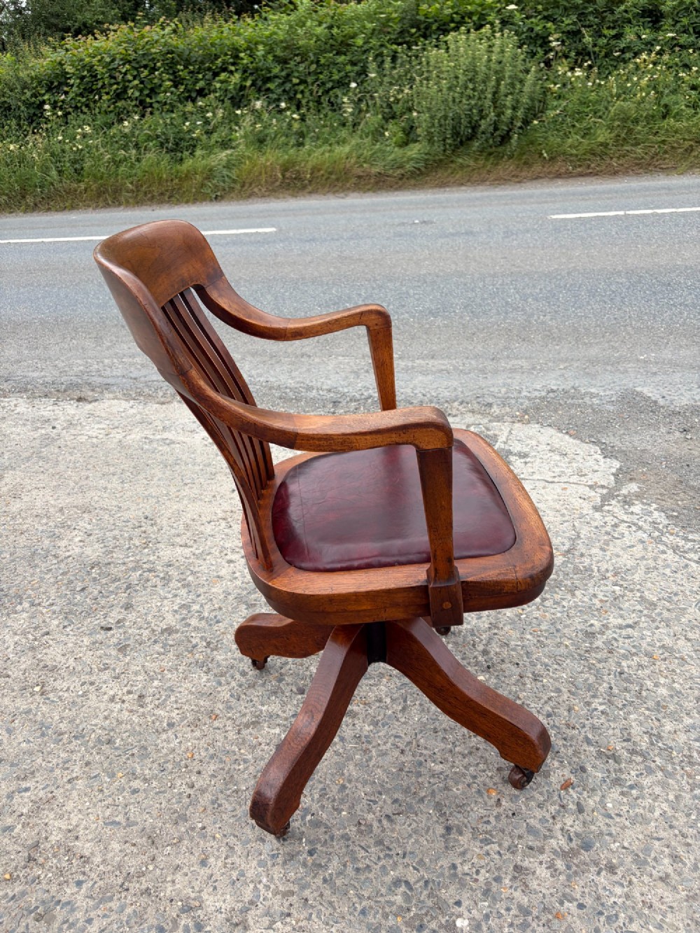 A 1920s solid oak swivel desk chair featuring a slatted backrest, integrated armrests, a reddish-brown leather seat cushion, and a four-star base on castors. View II