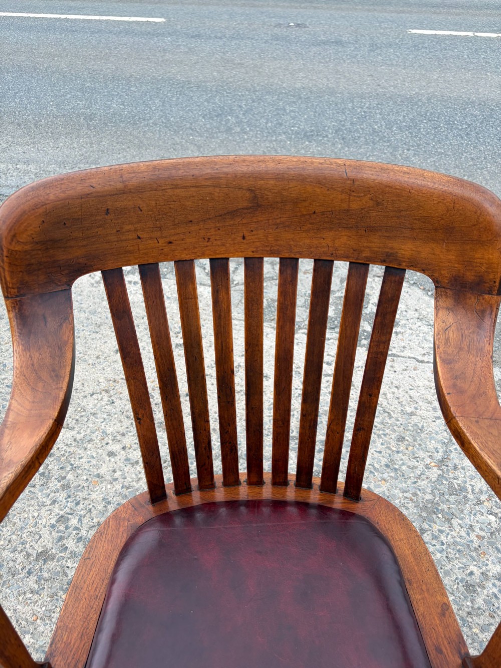A 1920s solid oak swivel desk chair featuring a slatted backrest, integrated armrests, a reddish-brown leather seat cushion, and a four-star base on castors. View III