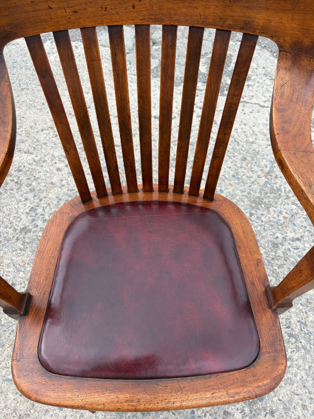 A 1920s solid oak swivel desk chair featuring a slatted backrest, integrated armrests, a reddish-brown leather seat cushion, and a four-star base on castors. View IV