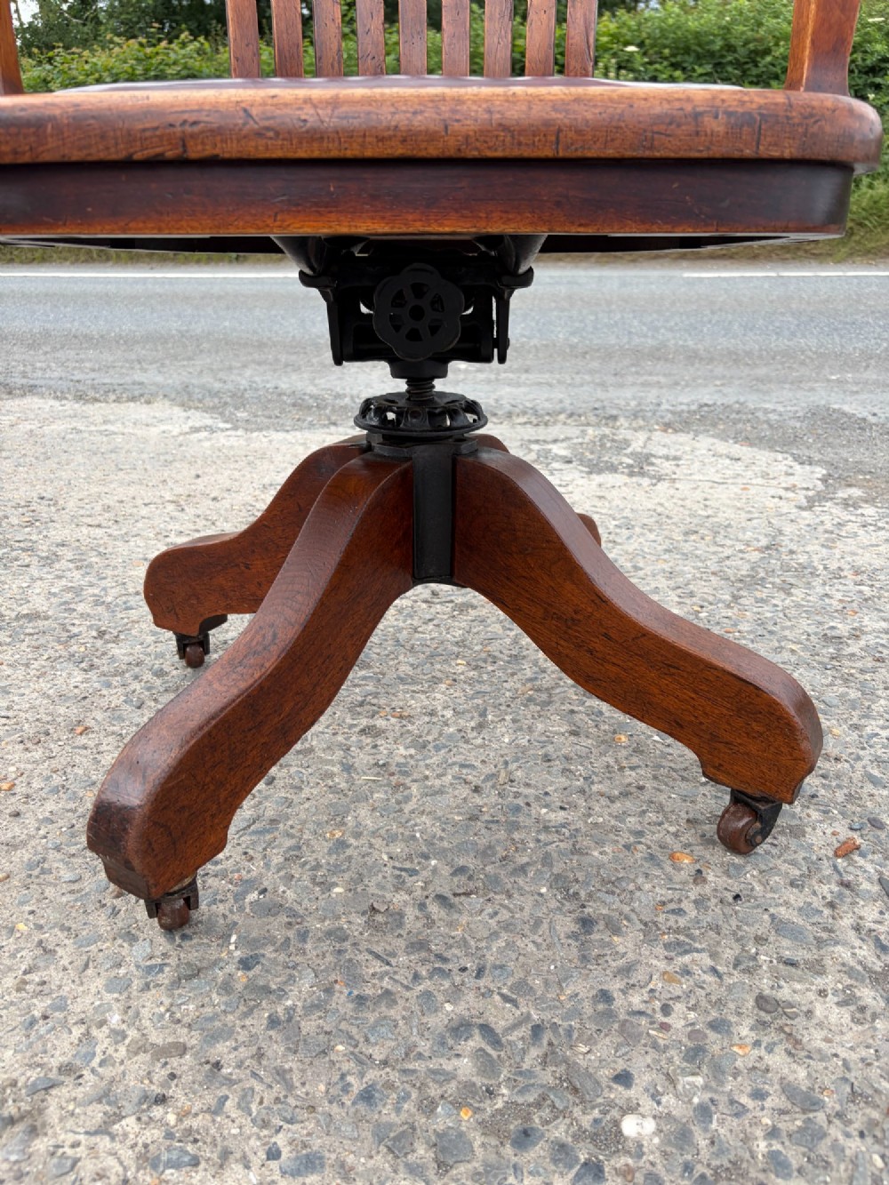 A 1920s solid oak swivel desk chair featuring a slatted backrest, integrated armrests, a reddish-brown leather seat cushion, and a four-star base on castors. View V