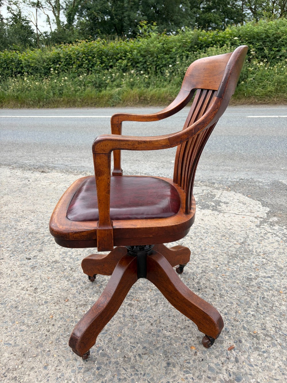 A 1920s solid oak swivel desk chair featuring a slatted backrest, integrated armrests, a reddish-brown leather seat cushion, and a four-star base on castors. View VI