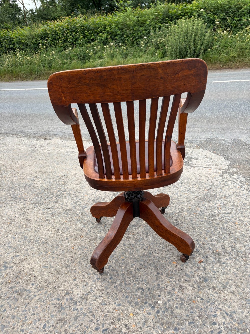 A 1920s solid oak swivel desk chair featuring a slatted backrest, integrated armrests, a reddish-brown leather seat cushion, and a four-star base on castors. View VII