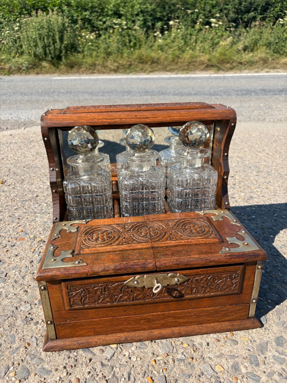 A late Victorian carved oak tantalus set featuring brass corner mounts, a front compartment with carved scrollwork, and three cut glass decanters backed by a mirror.