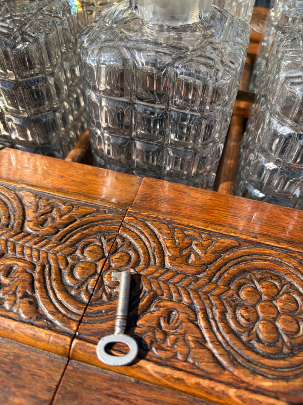 A late Victorian carved oak tantalus set featuring brass corner mounts, a front compartment with carved scrollwork, and three cut glass decanters backed by a mirror. View II