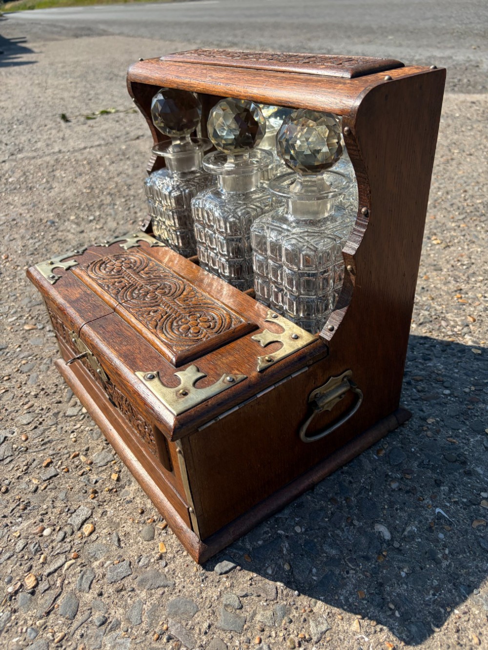 A late Victorian carved oak tantalus set featuring brass corner mounts, a front compartment with carved scrollwork, and three cut glass decanters backed by a mirror. View V