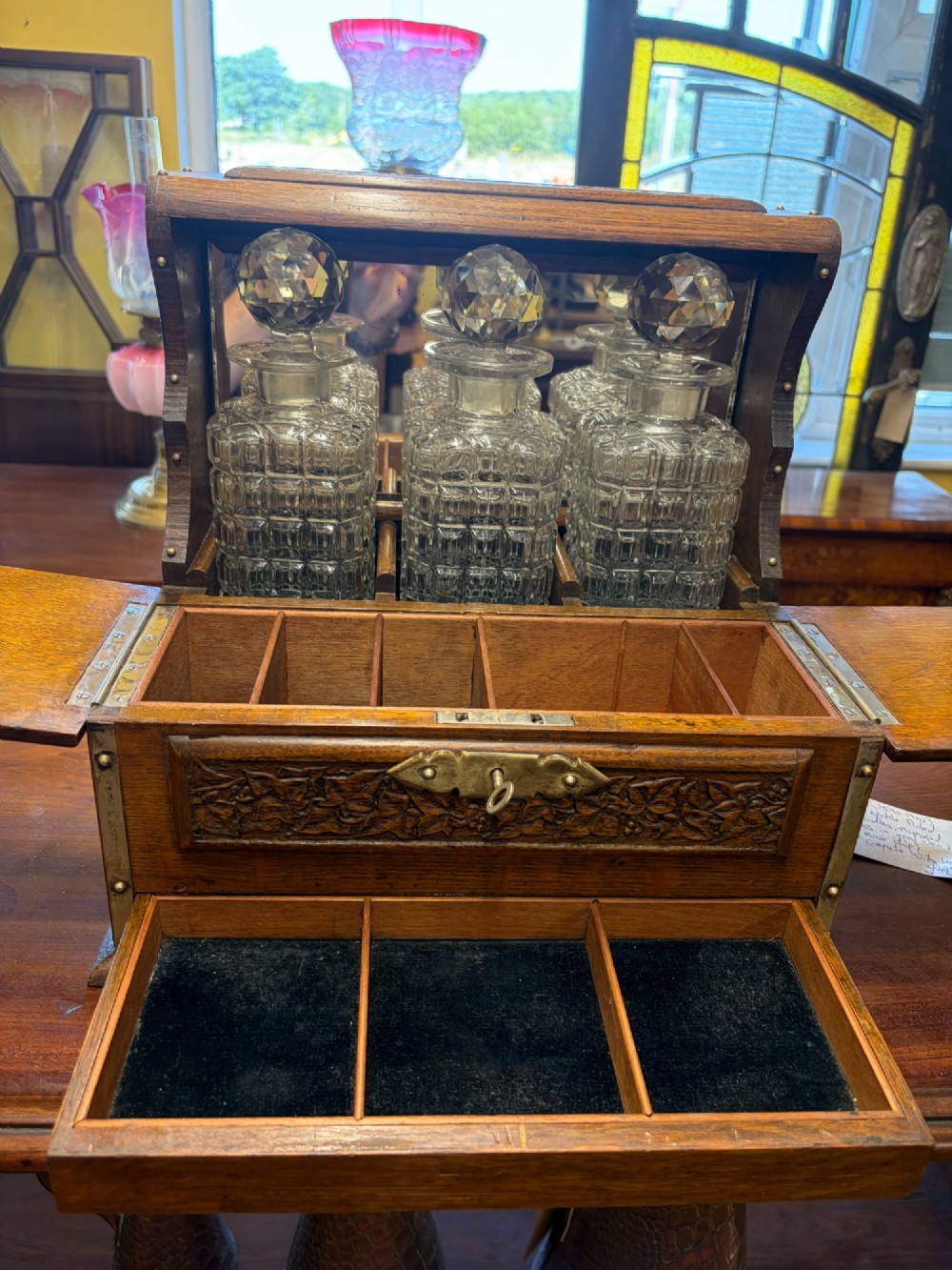A late Victorian carved oak tantalus set featuring brass corner mounts, a front compartment with carved scrollwork, and three cut glass decanters backed by a mirror. View VIII