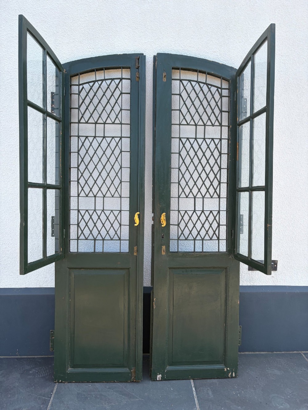 A pair of dark green painted pine double doors featuring arched tops, iron lattice grilles, and opening glazed window frames above solid lower timber panels.