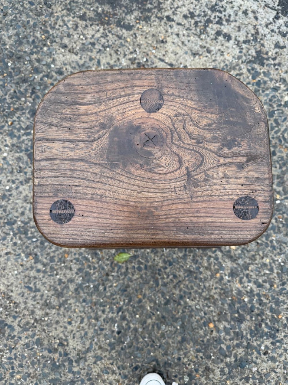 A three-legged dark brown timber artisan stool featuring exposed wedged tenons on the seat surface and triangular cross stretchers. View III