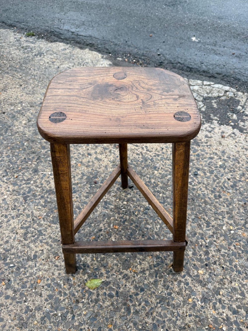 A three-legged dark brown timber artisan stool featuring exposed wedged tenons on the seat surface and triangular cross stretchers.