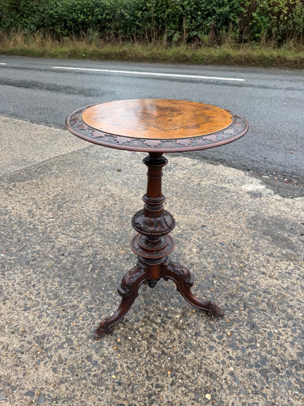A Victorian mahogany tripod side table with a circular top containing a burr wood inset, a carved border, a turned central support, and three carved splayed legs.