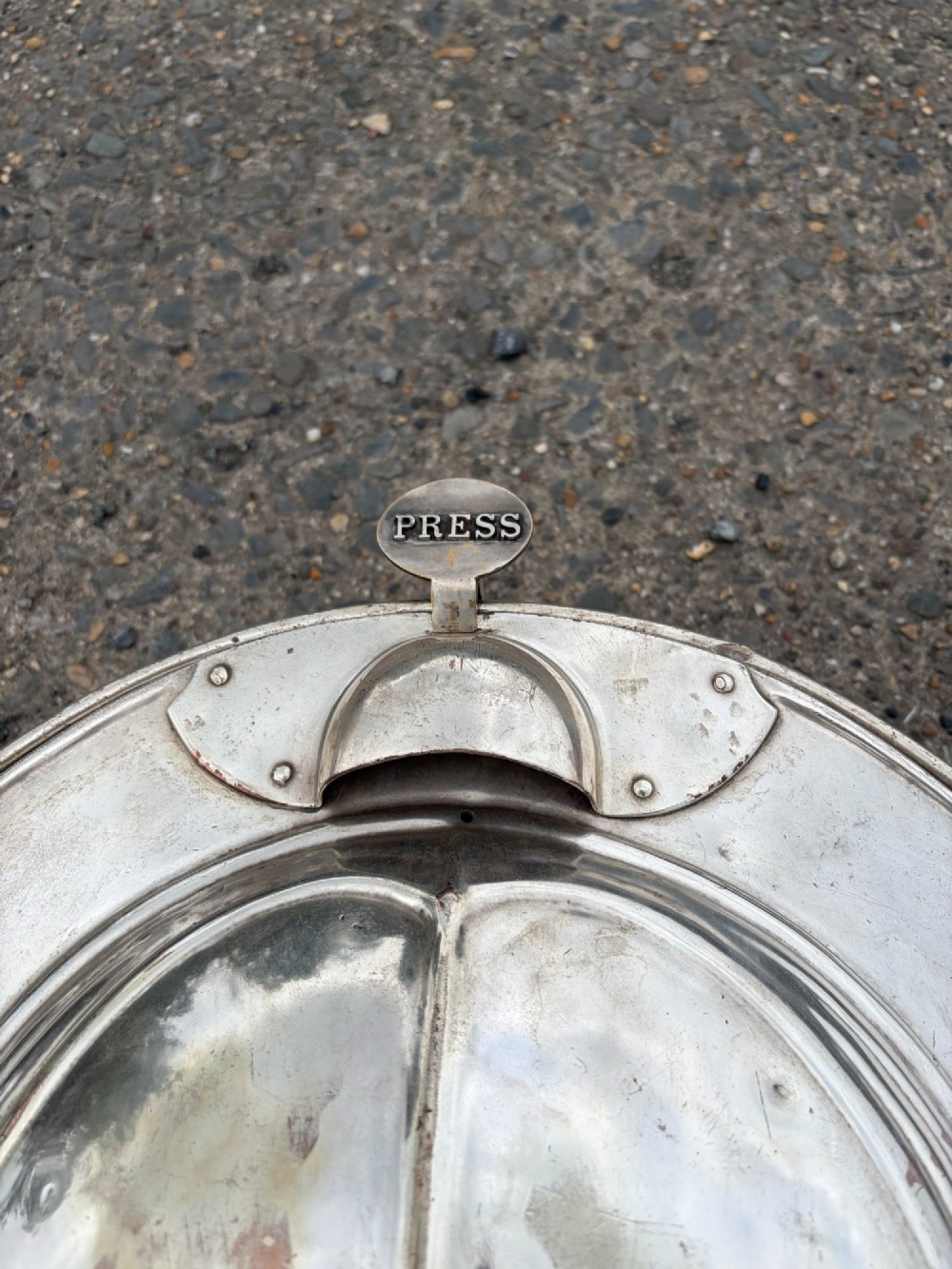 An oval silver-plated railway carriage folding sink featuring relief scrollwork, a press-button top mechanism, and exposed brass pipe fittings. View V