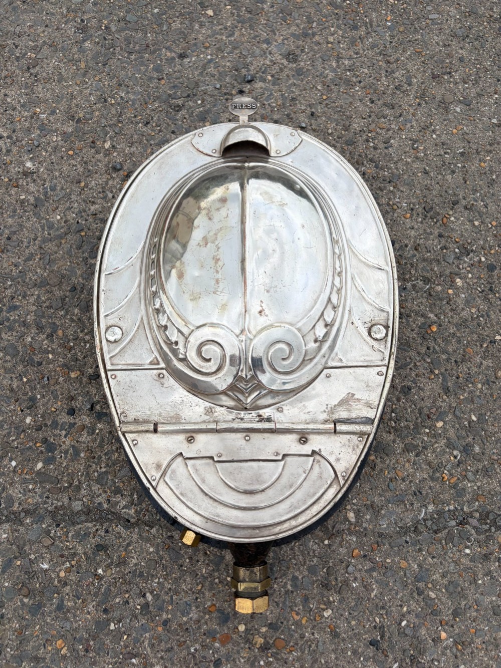 An oval silver-plated railway carriage folding sink featuring relief scrollwork, a press-button top mechanism, and exposed brass pipe fittings.