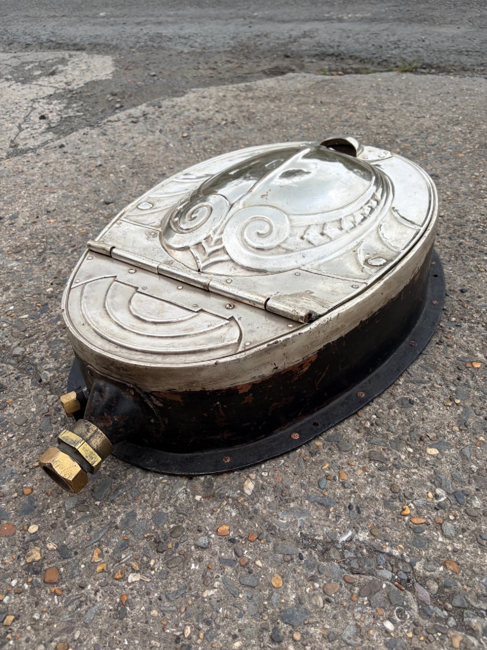 An oval silver-plated railway carriage folding sink featuring relief scrollwork, a press-button top mechanism, and exposed brass pipe fittings. View XII
