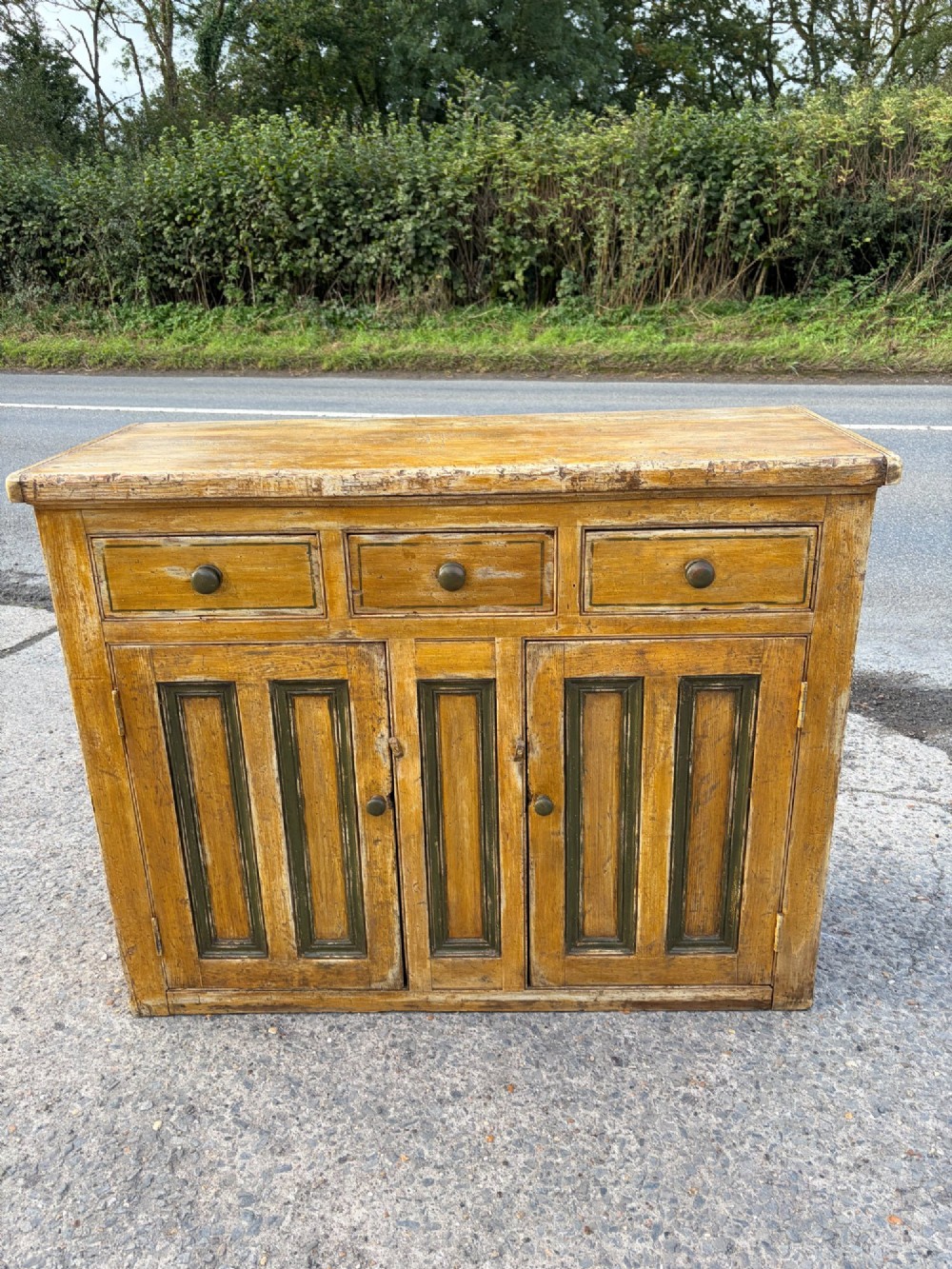 A late Victorian pine dresser base featuring a distressed ochre and green painted finish, three upper drawers, and recessed panel cupboard doors with wooden handles. View II