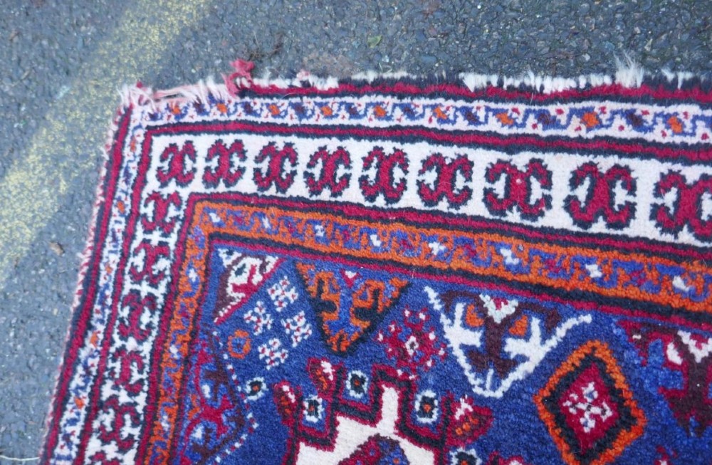 A rectangular hand-woven Persian wool carpet featuring a red stepped diamond centre, indigo blue spandrels, and geometric tribal borders. View V