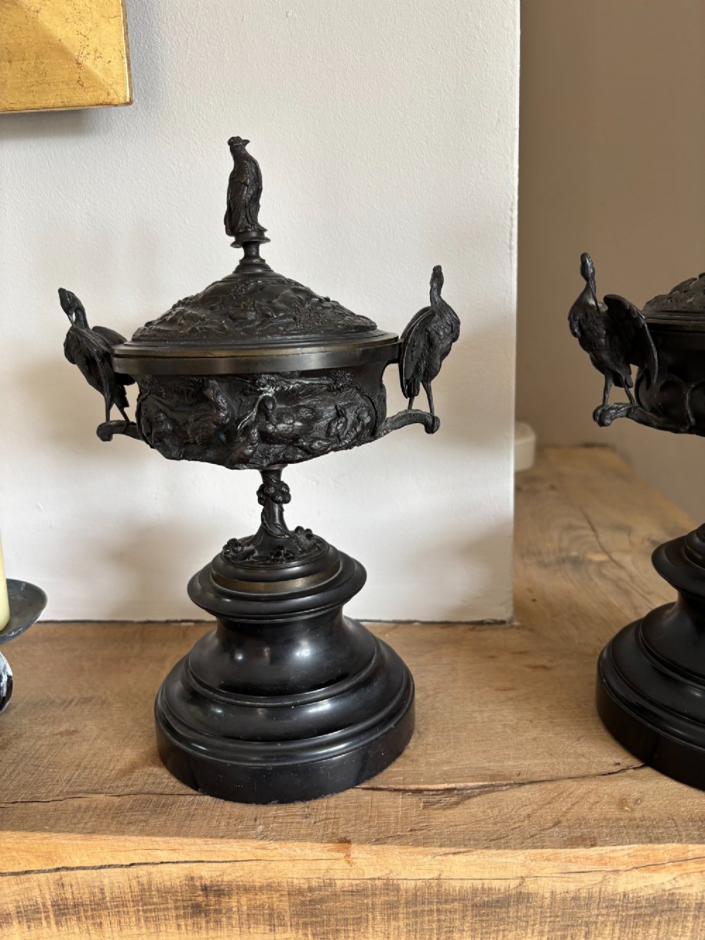 A pair of dark patinated bronze covered urns featuring relief-sculpted avian motifs and stork-shaped handles and finials on tiered bases. View II