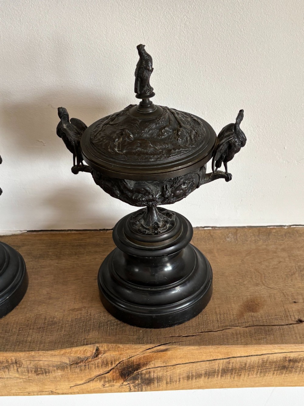 A pair of dark patinated bronze covered urns featuring relief-sculpted avian motifs and stork-shaped handles and finials on tiered bases. View X