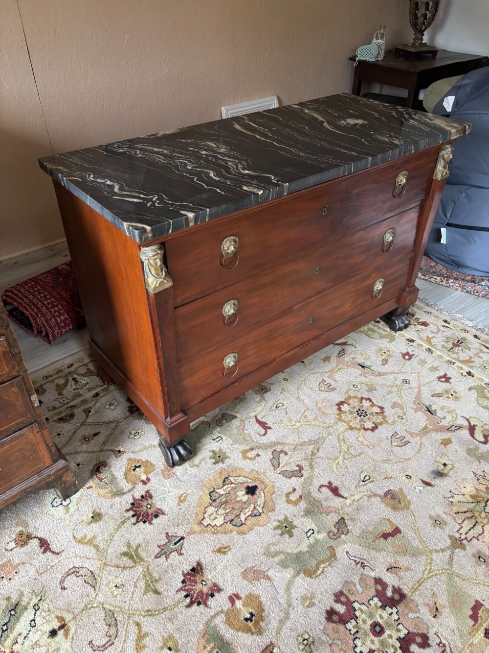 An early nineteenth-century French Empire mahogany commode featuring three drawers with brass lion-mask ring handles, gilt female caryatid mounts at the upper corners, a dark veined marble top, and sculpted paw feet.