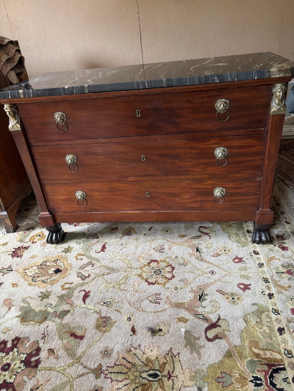 An early nineteenth-century French Empire mahogany commode featuring three drawers with brass lion-mask ring handles, gilt female caryatid mounts at the upper corners, a dark veined marble top, and sculpted paw feet. View IV