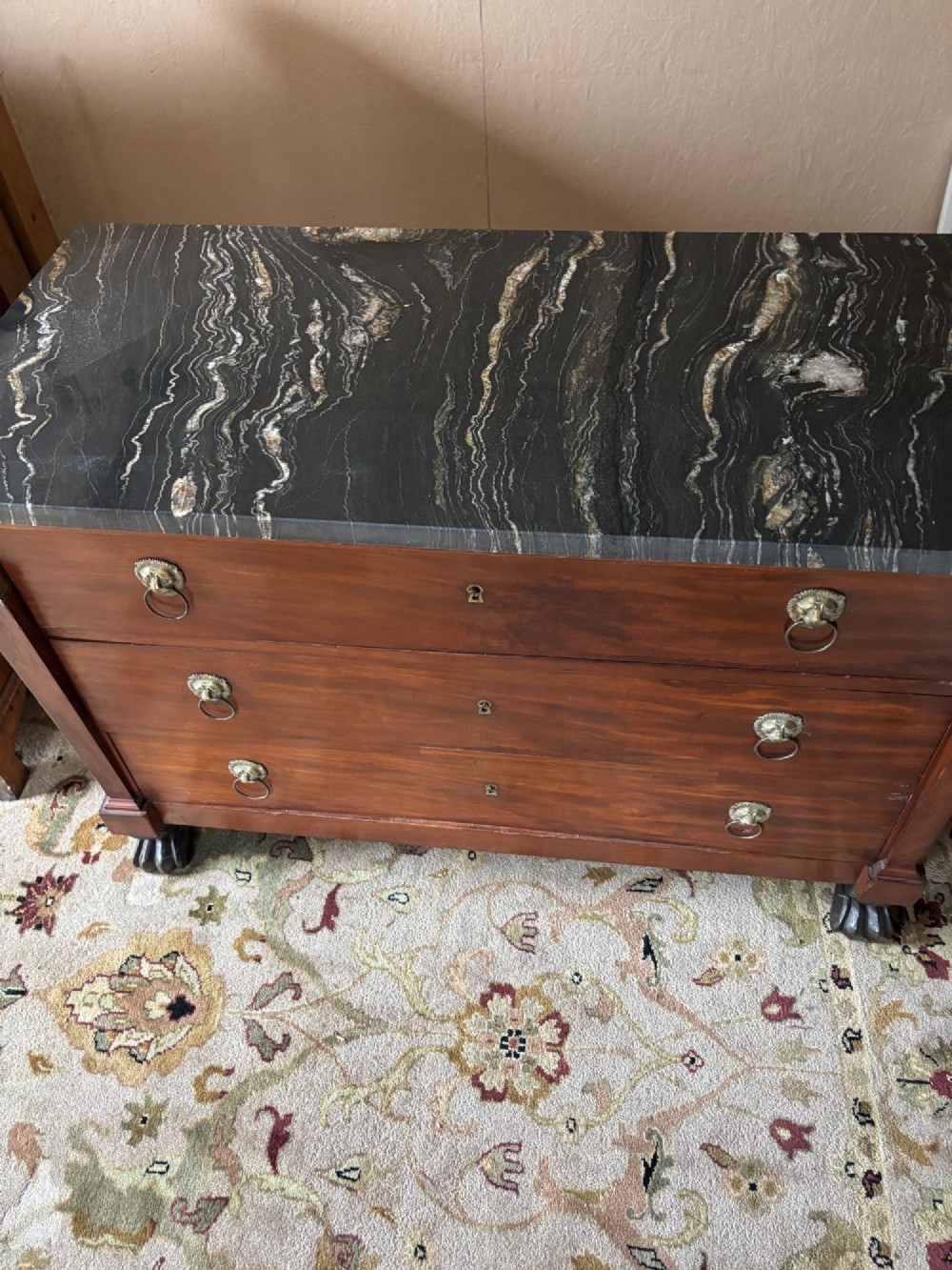 An early nineteenth-century French Empire mahogany commode featuring three drawers with brass lion-mask ring handles, gilt female caryatid mounts at the upper corners, a dark veined marble top, and sculpted paw feet. View VI